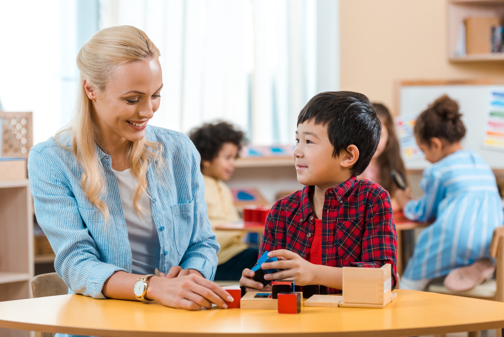 Selective focus of smiling teacher and kid playing building blocks with children at background in